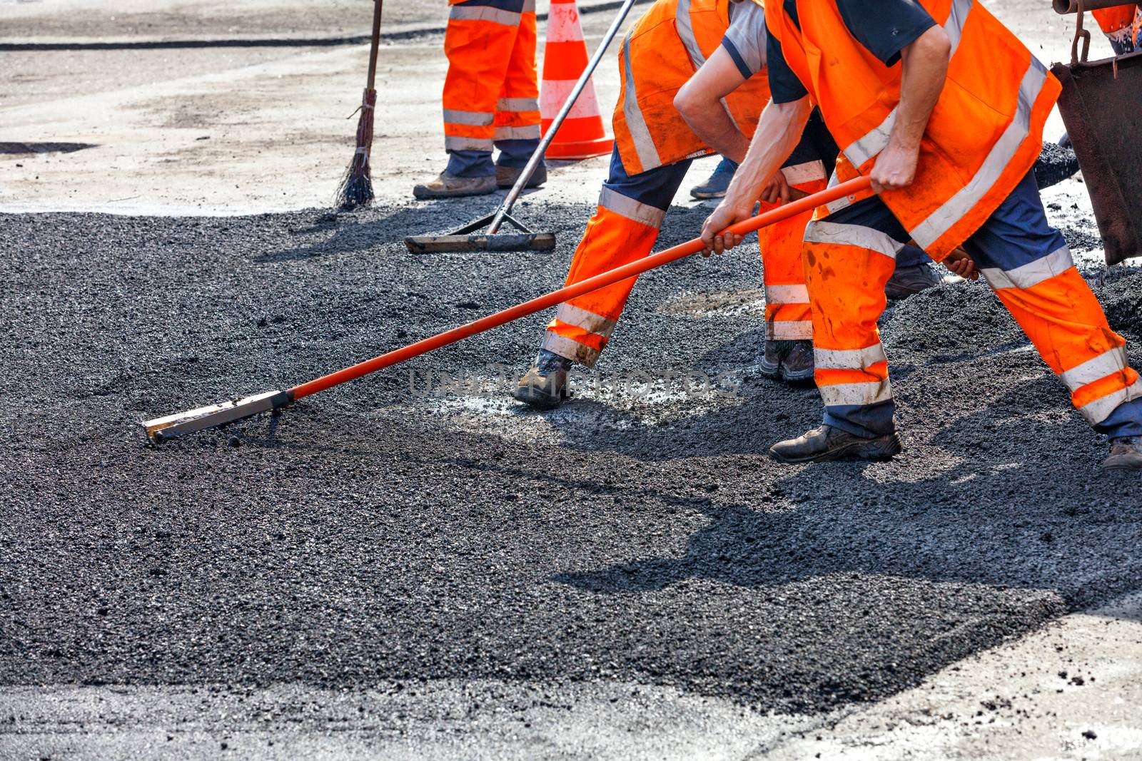 A working group of road workers in orange overalls renews a section of the road with fresh hot asphalt and smooths it out for repair with metal levels.