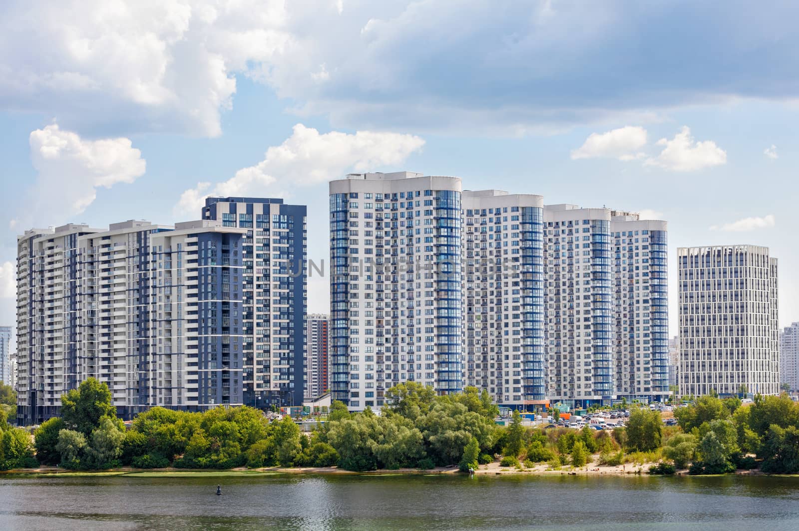 The facades of new residential high-rise buildings on the banks of the river and against the blue cloudy sky. by Sergii