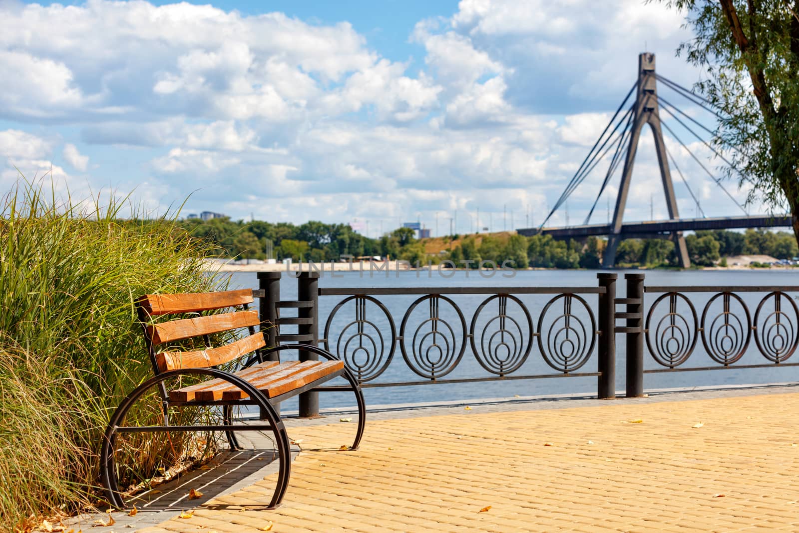 A beautiful wooden bench on the Dnipro embankment in Kyiv. Sidewalk yellow tiles against the backdrop of sunlight on a bright summer day and the blurred north bridge over the river.