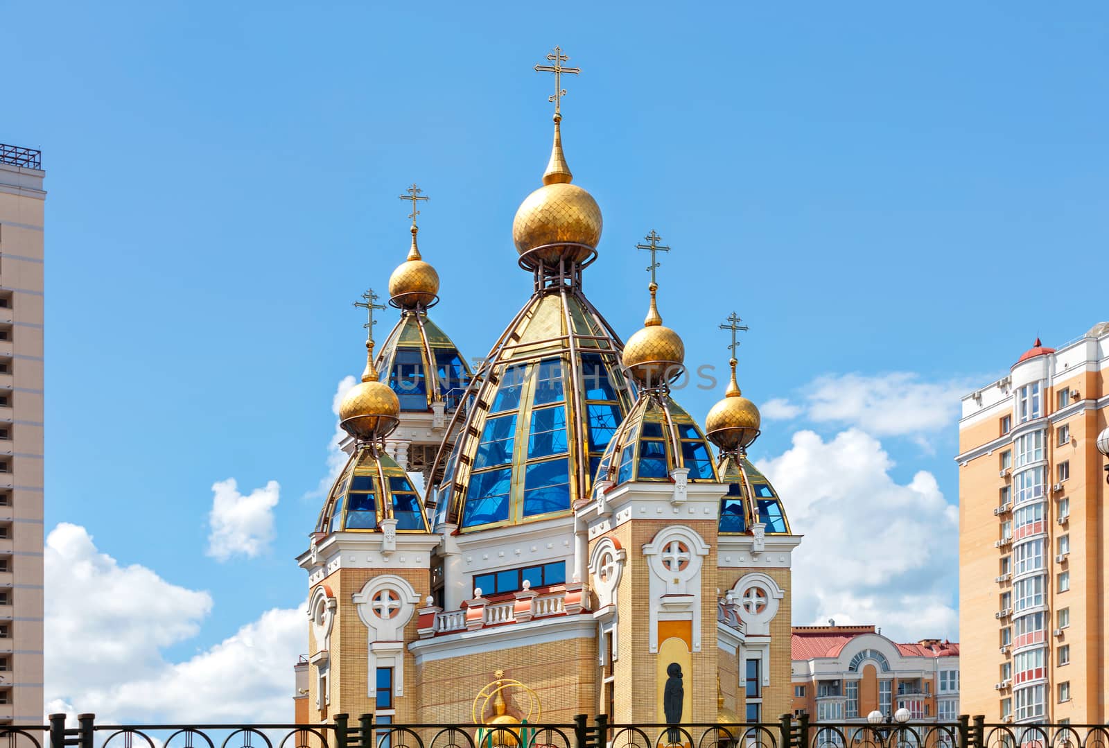 Golden domes of a Christian church on glass roof facades among an urban residential area against a blue cloudy sky.