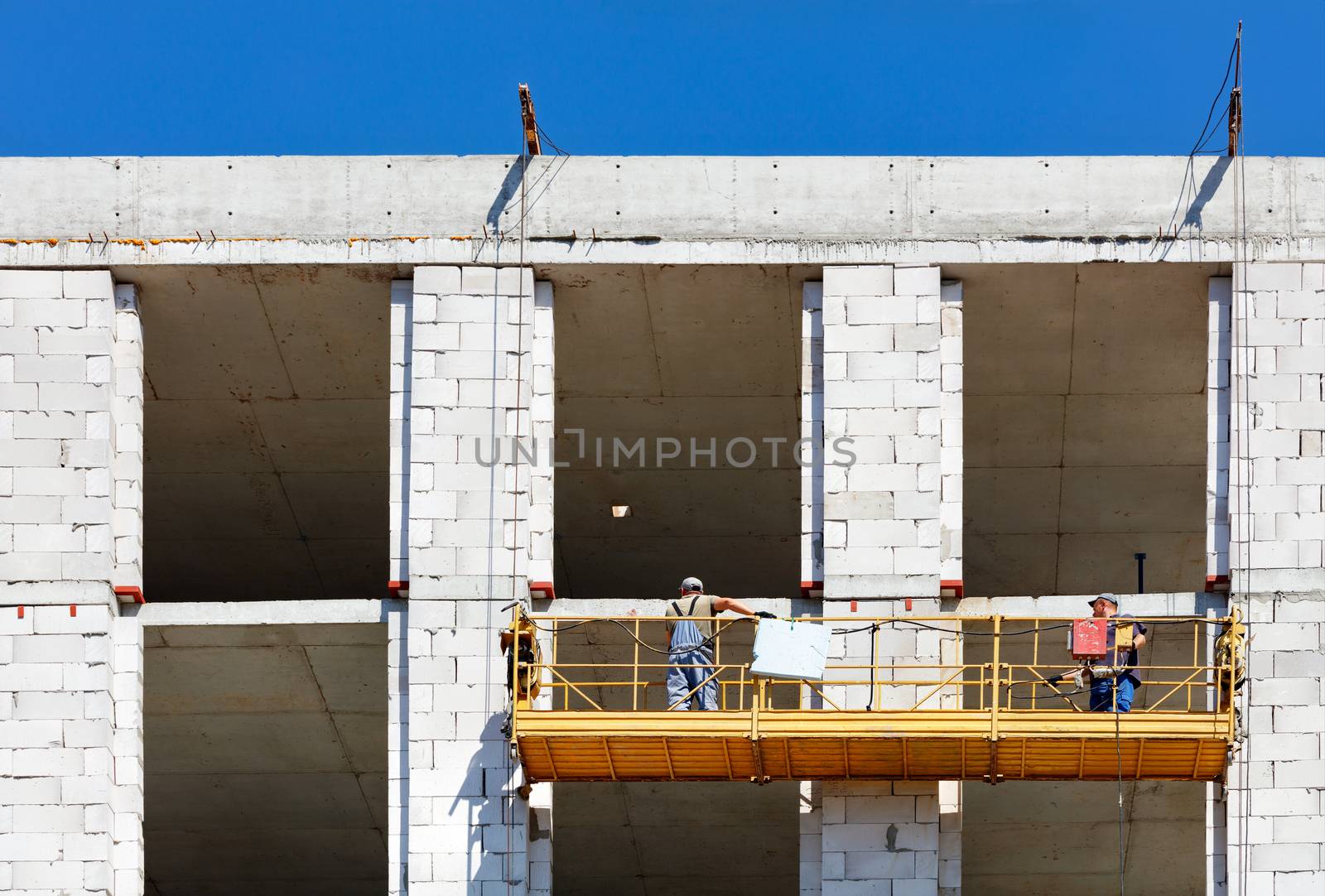 Construction workers climb on a cradle along the facade of a building under construction. by Sergii