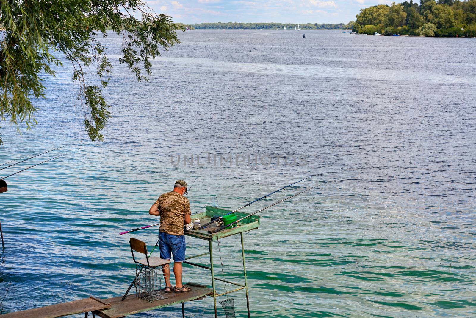 A fisherman catches fish with fishing rods on the river bank, standing on a wooden platform. by Sergii