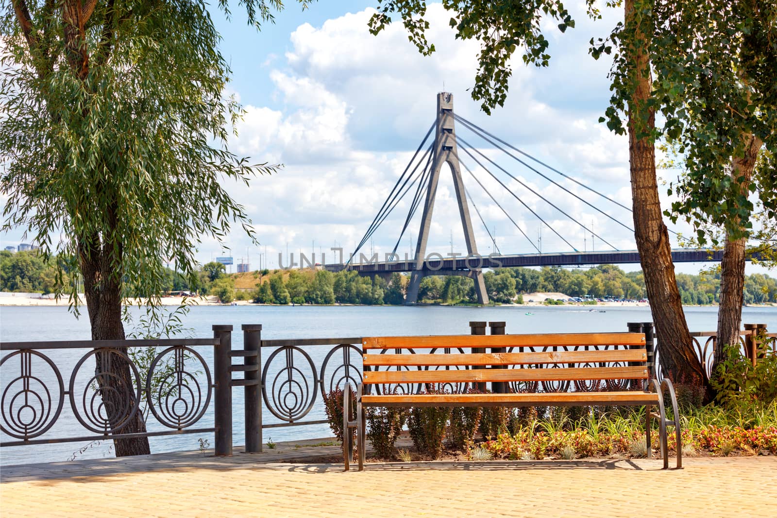 Summer landscape with a wooden bench on the Dnipro embankment against the background of the Northern bridge over the river in blur. by Sergii