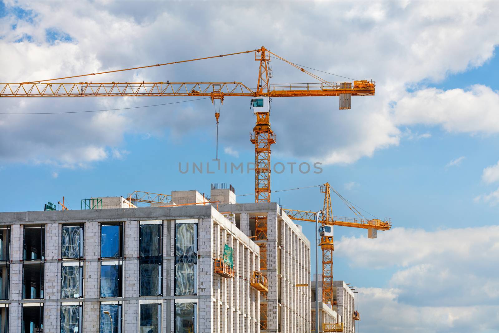 Tower cranes work on the construction site of a modern multi-storey residential building against a blue slightly cloudy sky, copy space.