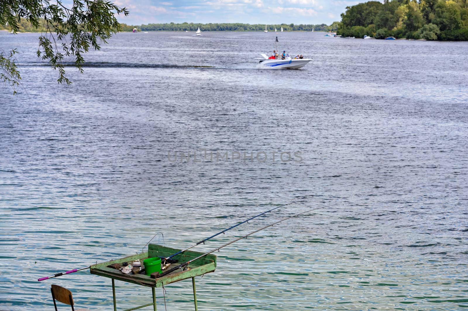 Summer landscape on the river bank with fishing rods and tackle against the backdrop of a motor boat and the opposite bank in blur, copy space.