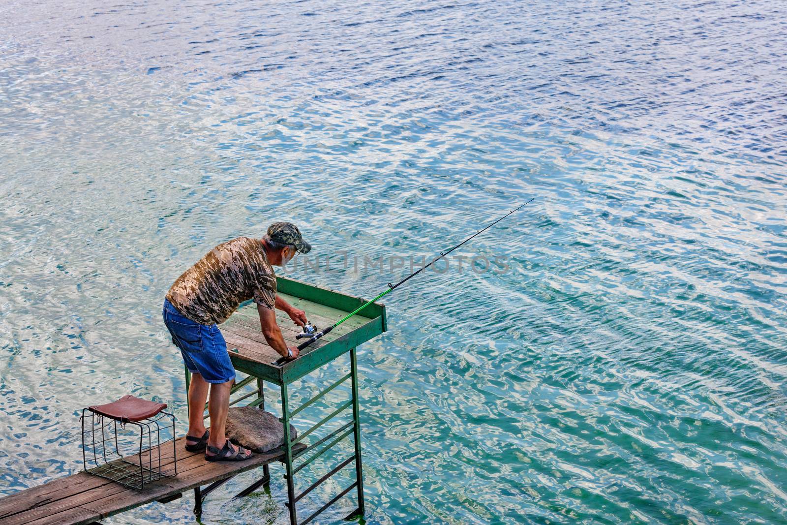 A fisherman in blue denim shorts is catching a spinning rod on the river bank on a summer morning, standing on a wooden platform against the backdrop of clean transparent water, copy space.