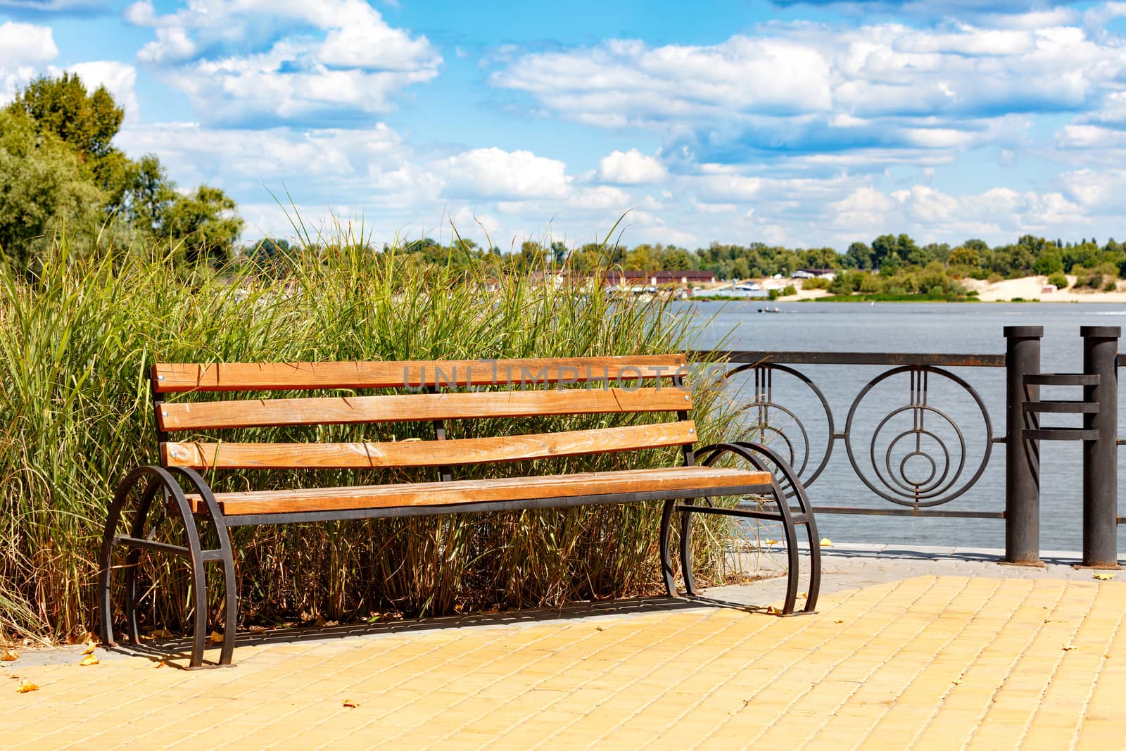 Wooden bench on the cobbled sidewalk of the Dnipro embankment against the backdrop of a summer landscape and of tall grass, a wide river of light cloudy sky, copy space.