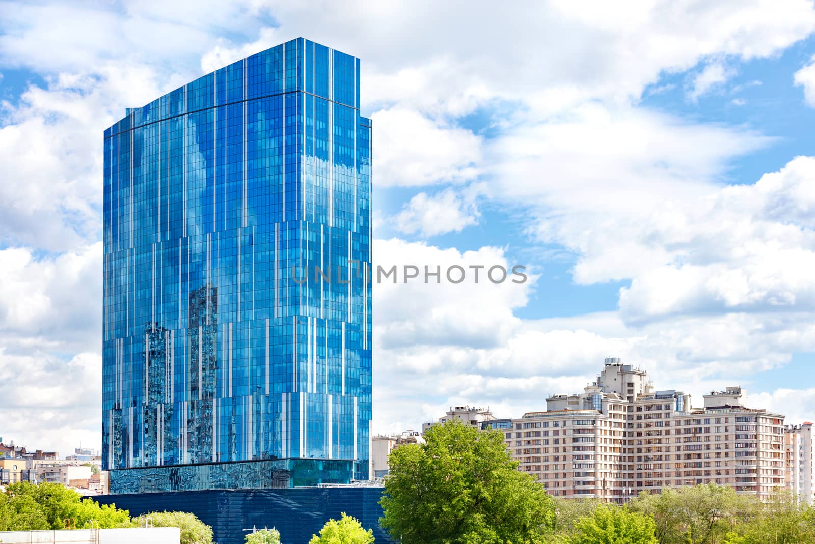 The beautiful glass facade of the modern building reflects the blue sky and white clouds in its windows against the backdrop of the cityscape.