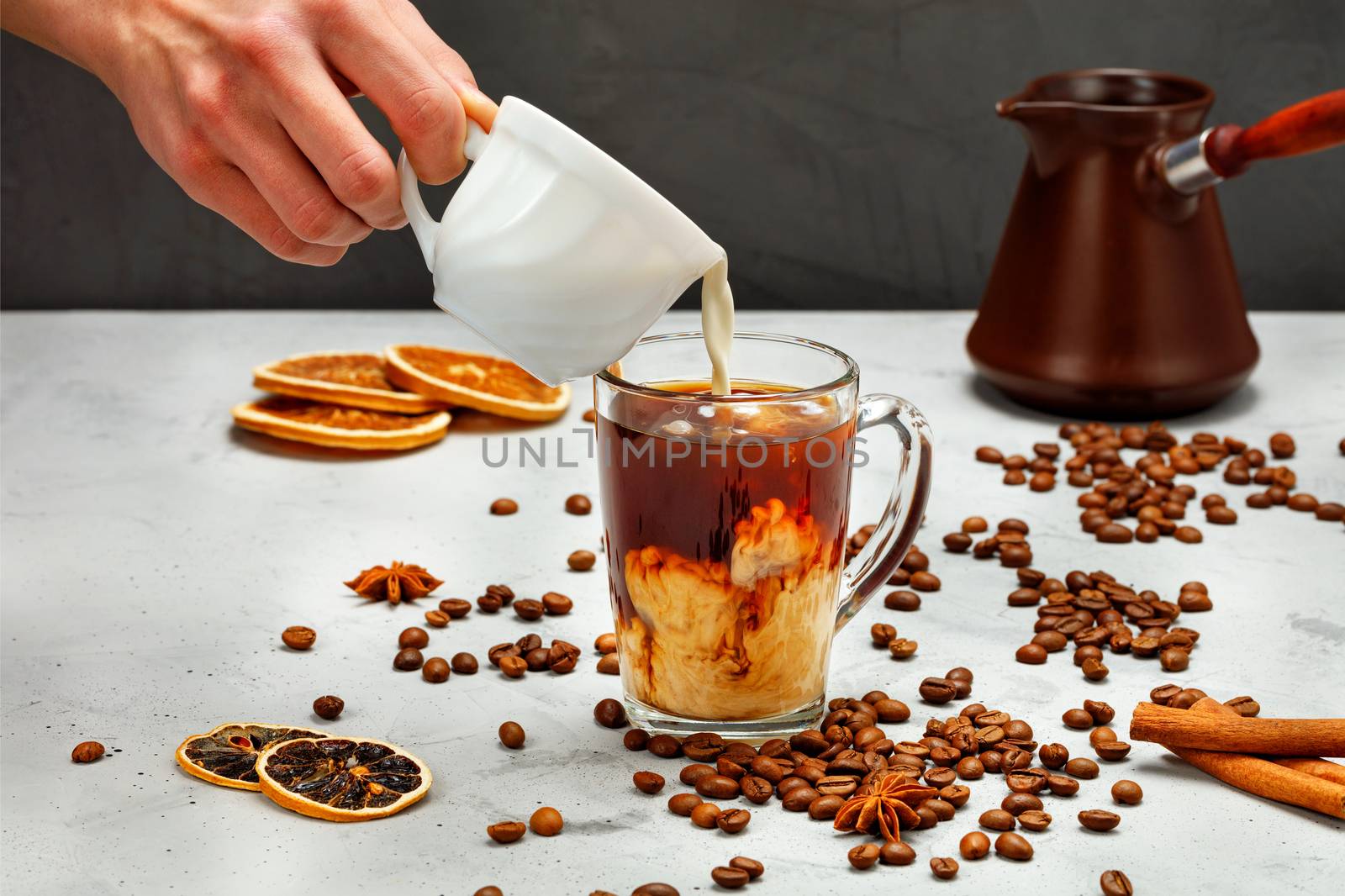 Male hand pouring cream from white cup into transparent glass with cold coffee, cold coffee concept on gray and black background, copy space.
