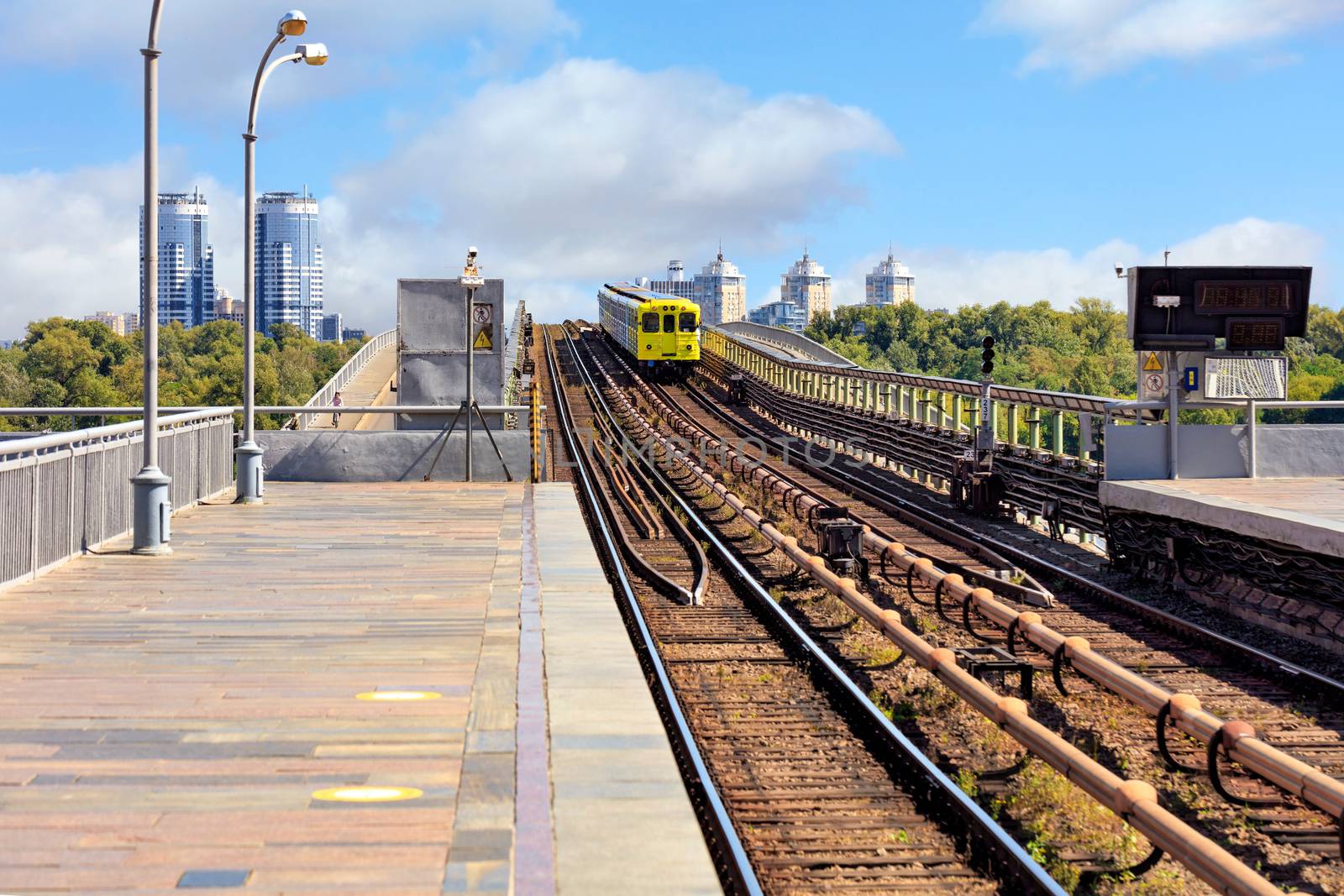 An open section of the railway track of a metro bridge in Kyiv, along which a lemon-colored subway train rushes against a backdrop of a cityscape and a blue sky. Foreground in a slight blur for copy space.