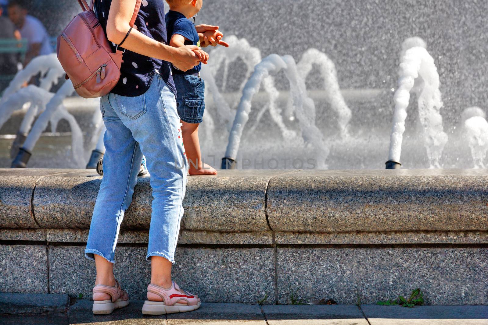 A mother with a small child stands at the city fountain in the summer heat to freshen up. by Sergii