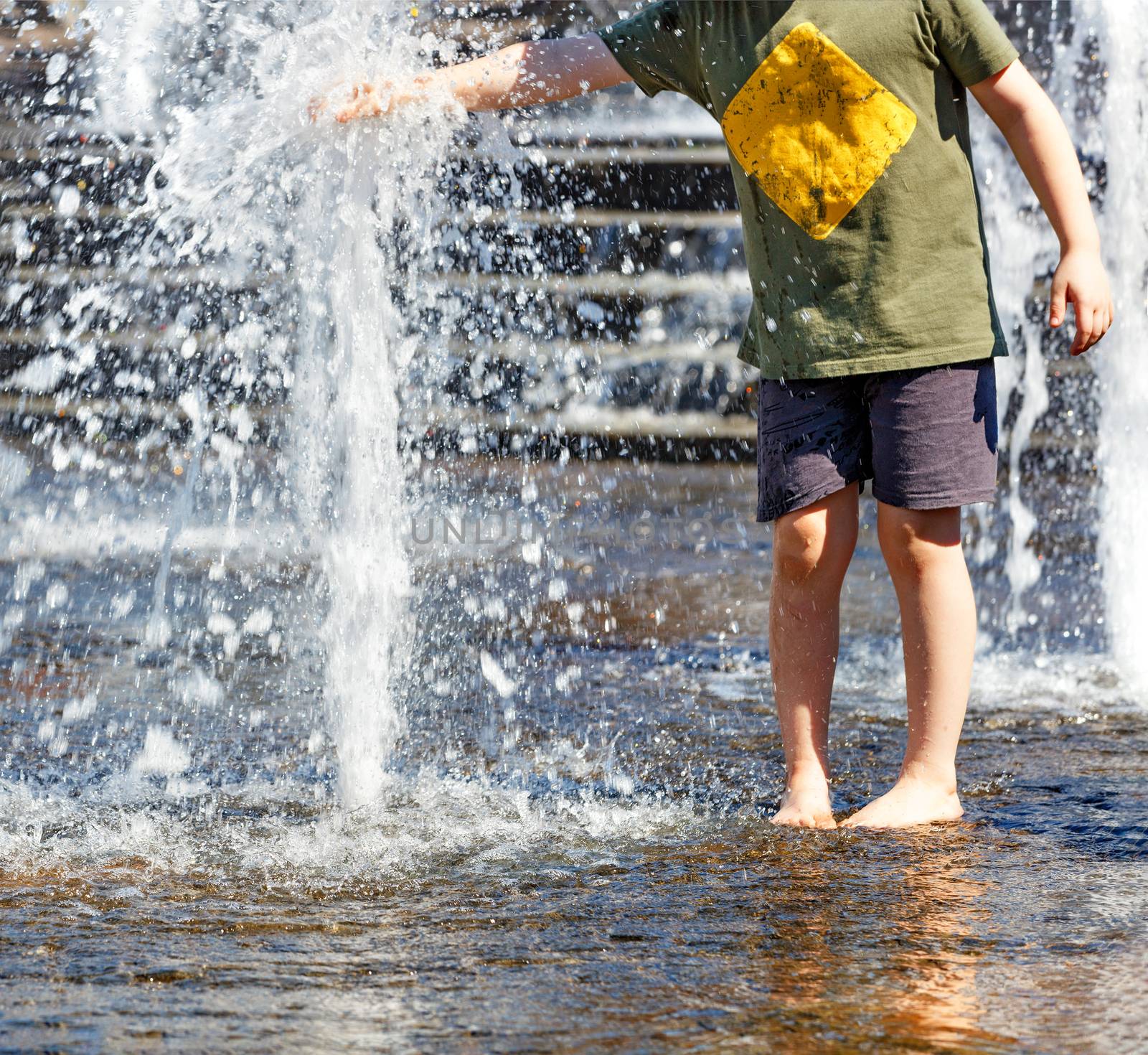 A teenager walks barefoot by the city fountain and tries to grab a powerful stream of water with his hand on a hot summer day.