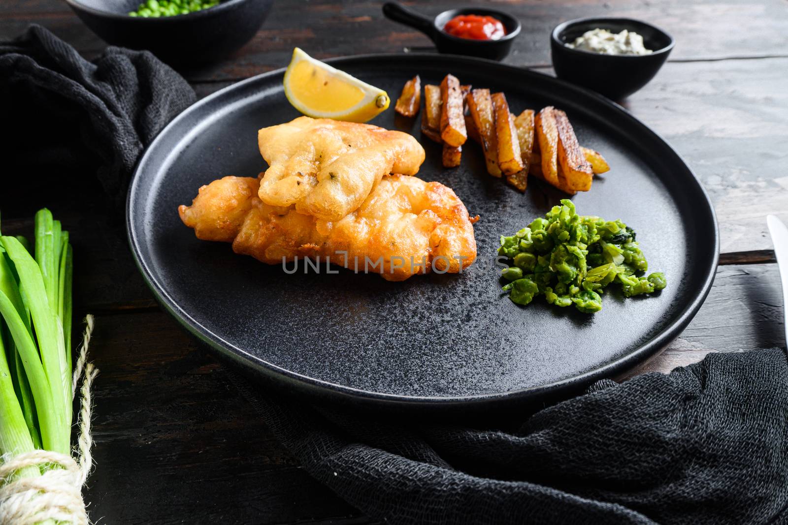 Fish and chips. Traditional british fast food hot dish consisting of fried fish, potato chips, mushy peas and tartare sauce. served on black plate over old dark wood table in pub side view.