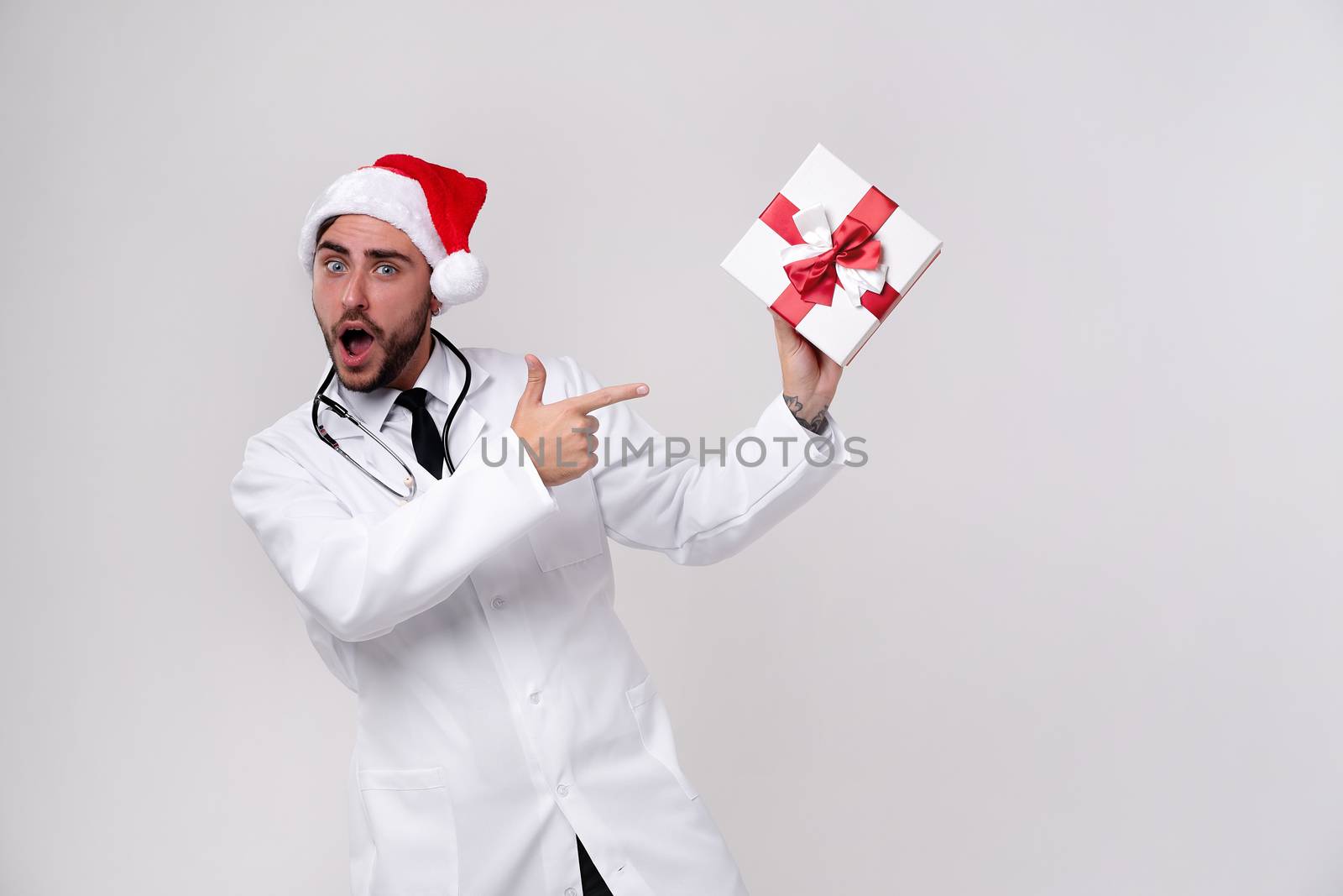 Young handsome doctor in white uniforme and Santa hat in studio white background smile Shows a finger at the gift box. showing thumbs up Medical student Christmas New Year Holiday Medicine concept