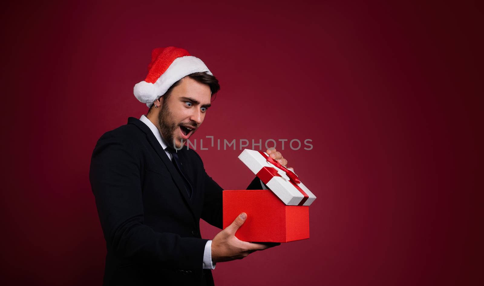 Surprised handsome caucasian guy in business suit and Santa hats stands on white background in studio and smilie opens red gift box in hand. Portrait business person with Christmas mood Holiday banner