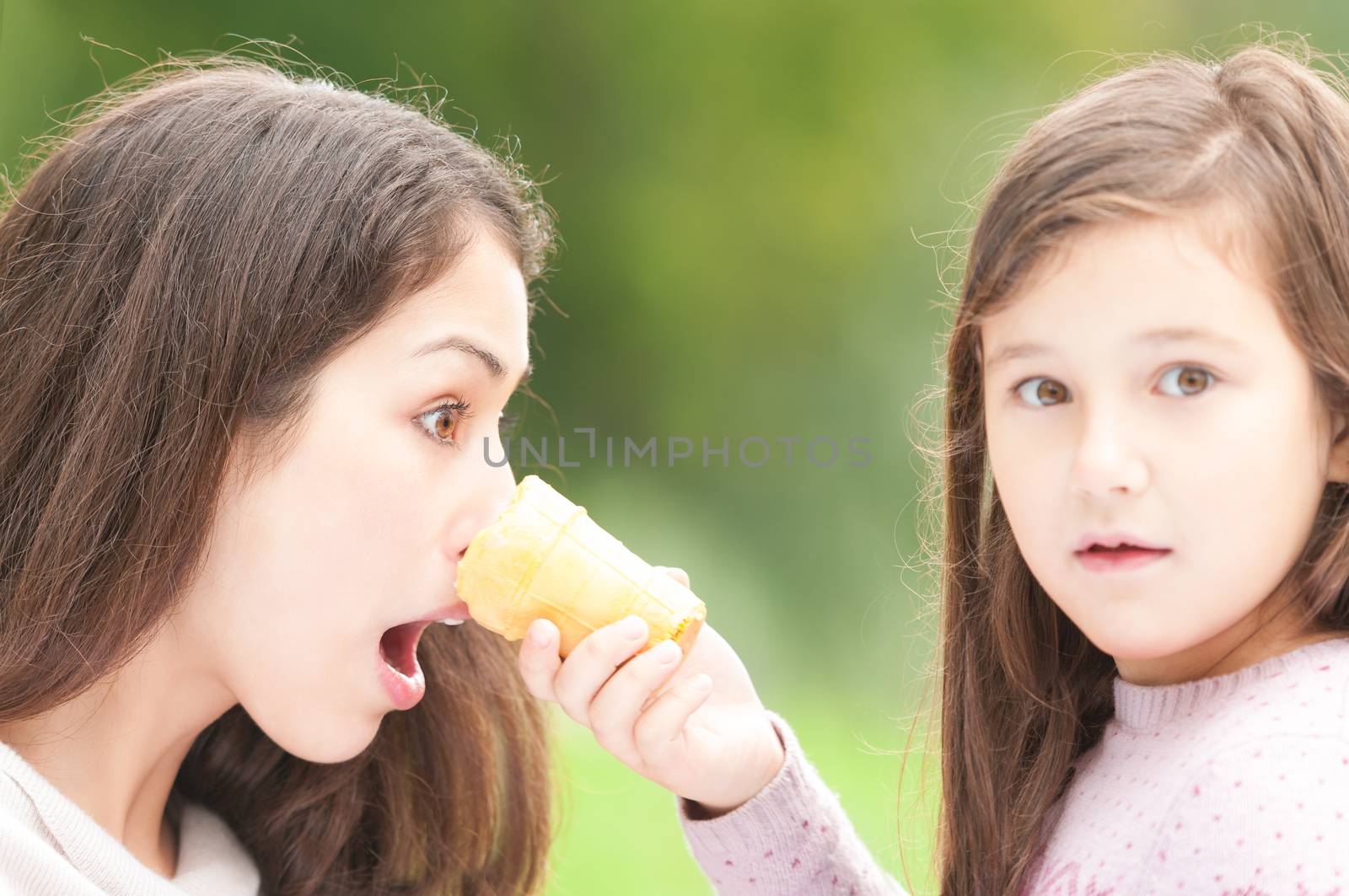 Small daughter pokes ice cream in face of her mother. Surprised facial expression of mother. Family outdoor activity in summer. Amazed emotion of woman and child.