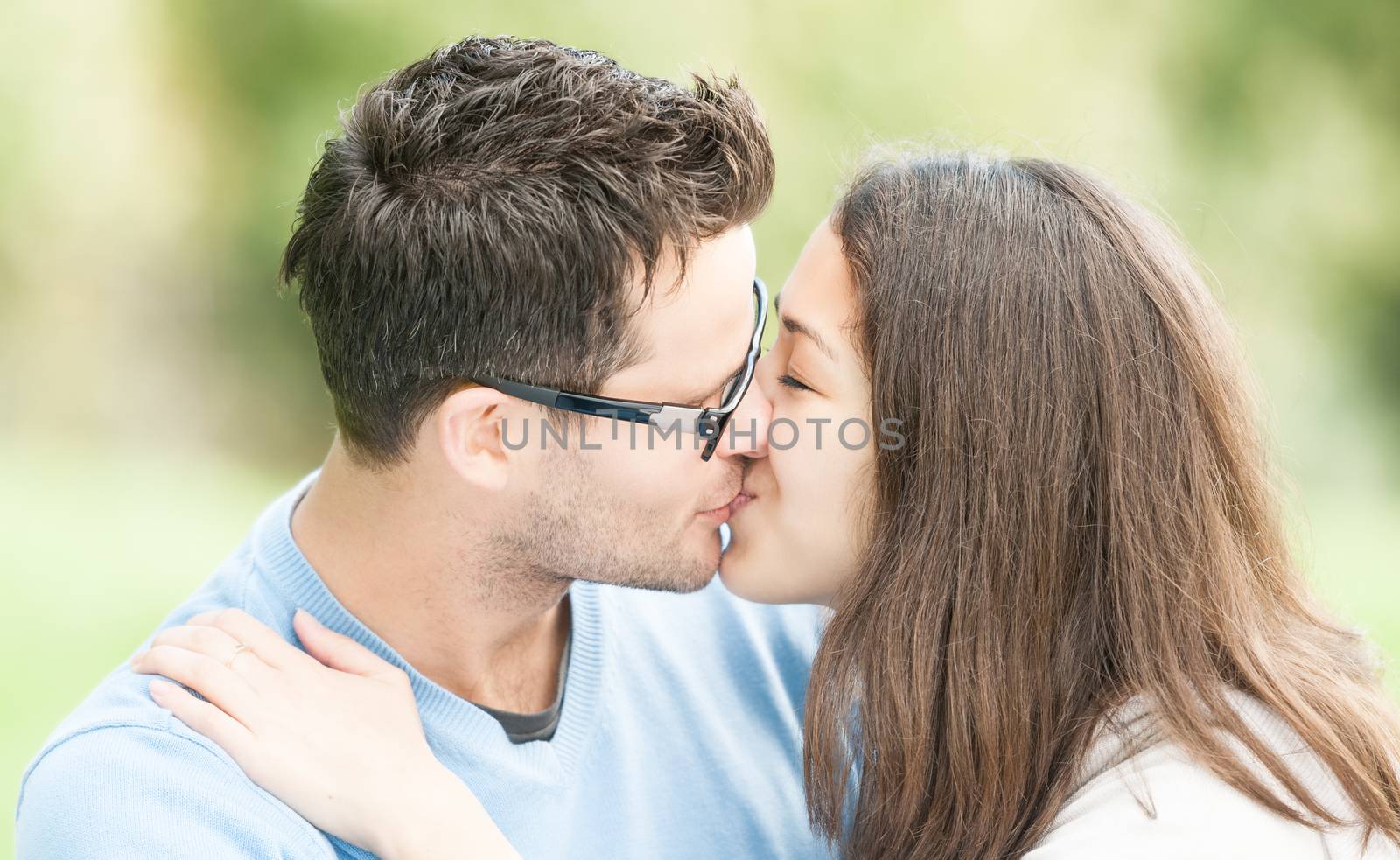 Handsome man in glasses and pretty woman kissing outdoors. Husband and wife giving kiss to each other in park. Happy family expressing feelings. Portrait of beautiful romantic couple in love.