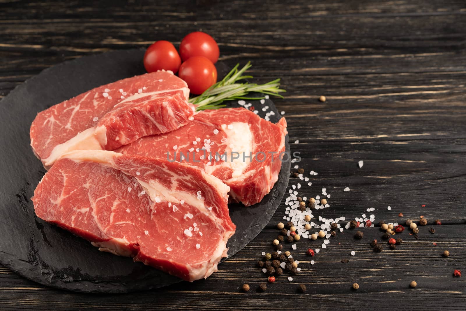 Three pieces of juicy raw beef with rosemary on a stone cutting board on a black wooden table background. Meat for barbecue or grill next to cherry tomatoes sprinkled with pepper and salt seasoning