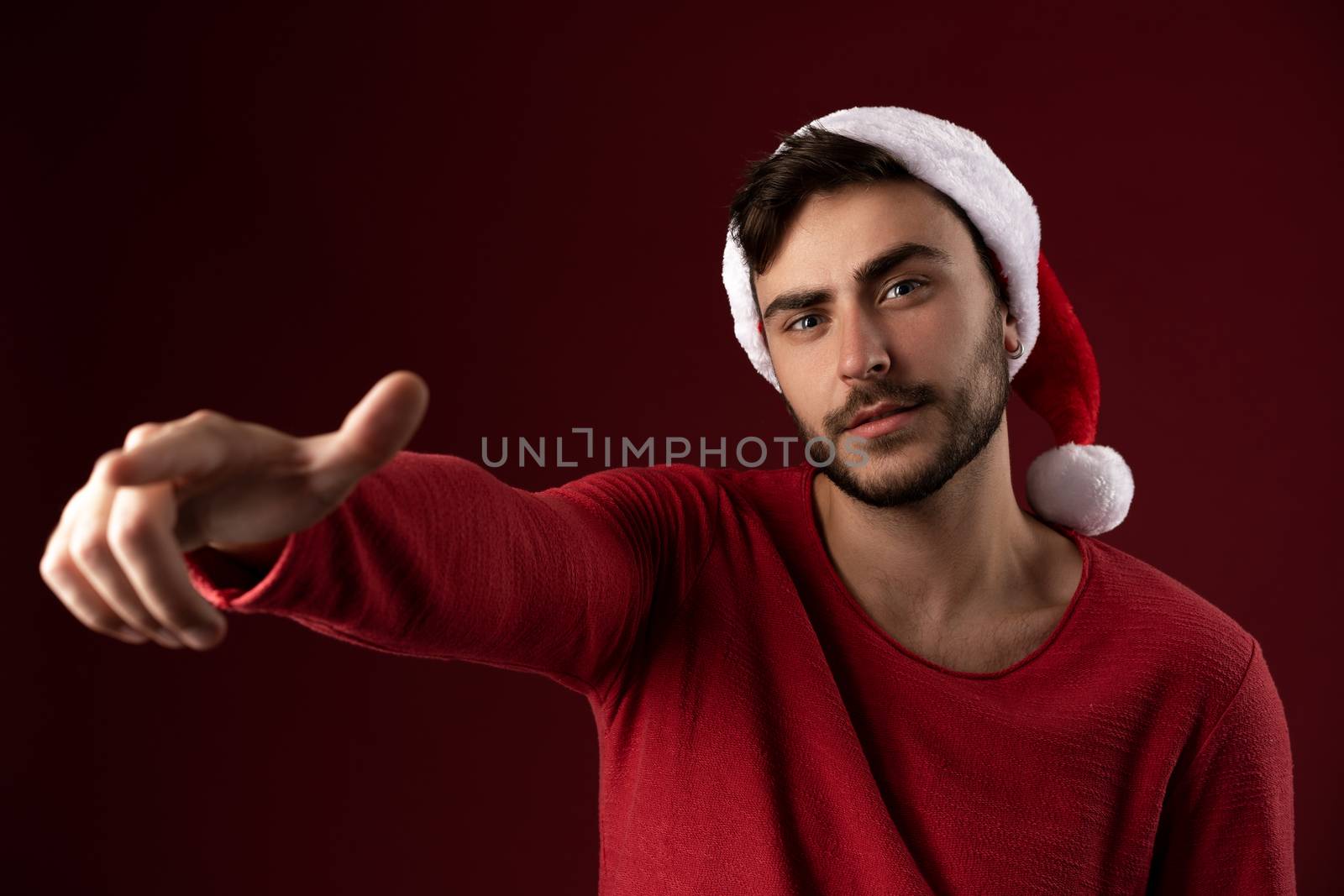 Young handsome caucasian guy in red sweater and Santa hats stands on red background in studio and Shows finger at the camera Close up portrait european guy with Christmas mood Holiday theme. New Year