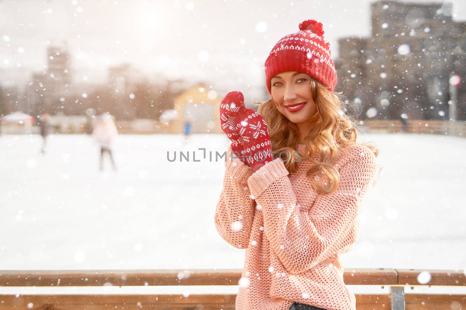 Beautiful lovely middle-aged girl curly hair warm peach sweater red knitted hat glove stands ice rink background Town Square Christmas mood lifestyle Happy holiday woman snowy day Winter leisure