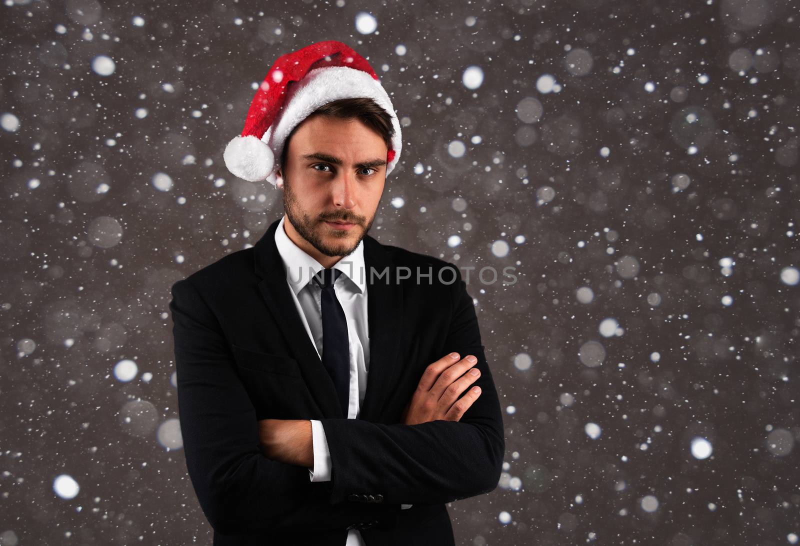 Young handsome caucasian guy in business suit and Santa hats stands on gray background in studio with serious face folded arms on chest Portrait business person with Christmas mood Holiday banner