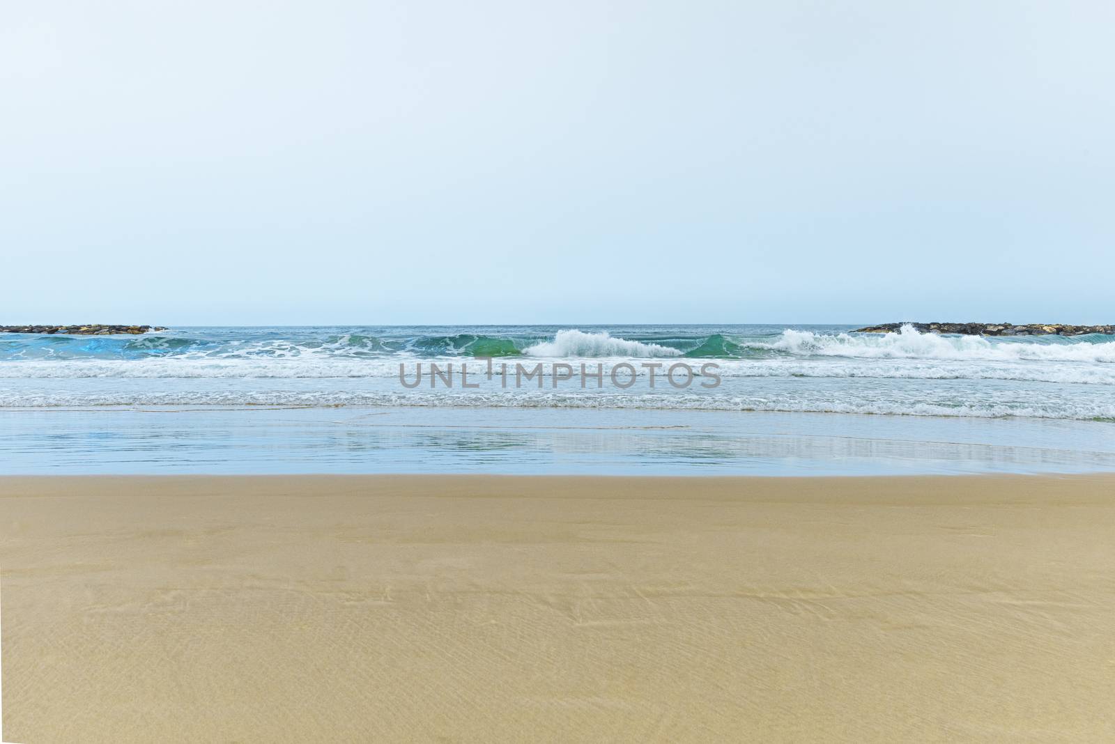 A sandy beach next to the ocean. A calm and quiet beach with deep turquoise clear water. Waves crashing on a breakwater. Soft, golden sea sand without a trace. The reflection of the sky on the water. Clear horizon line. High quality photo