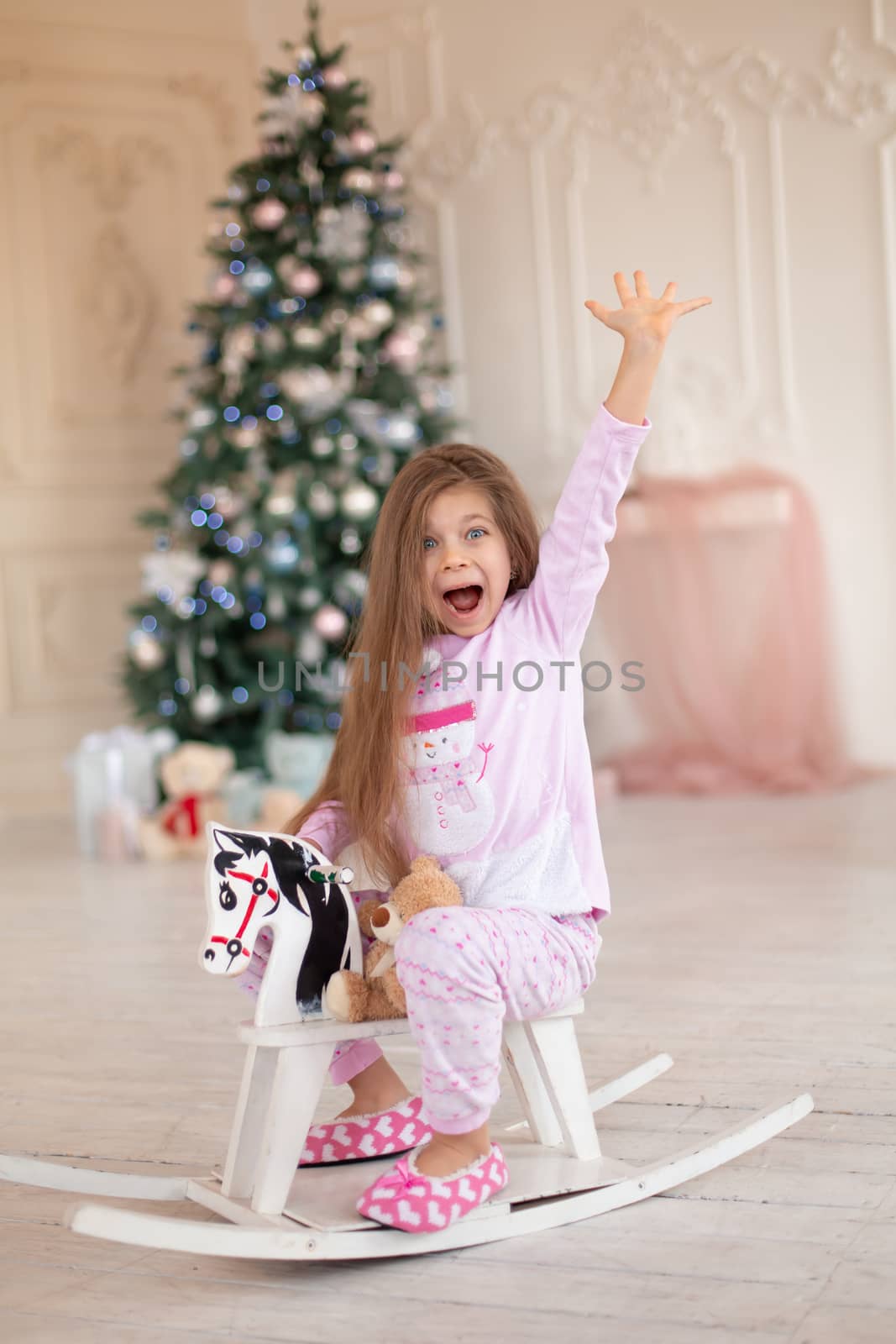 A beautiful little girl in pink pajamas rejoices in a wooden rocking horse, a gift from Santa for Christmas by Try_my_best
