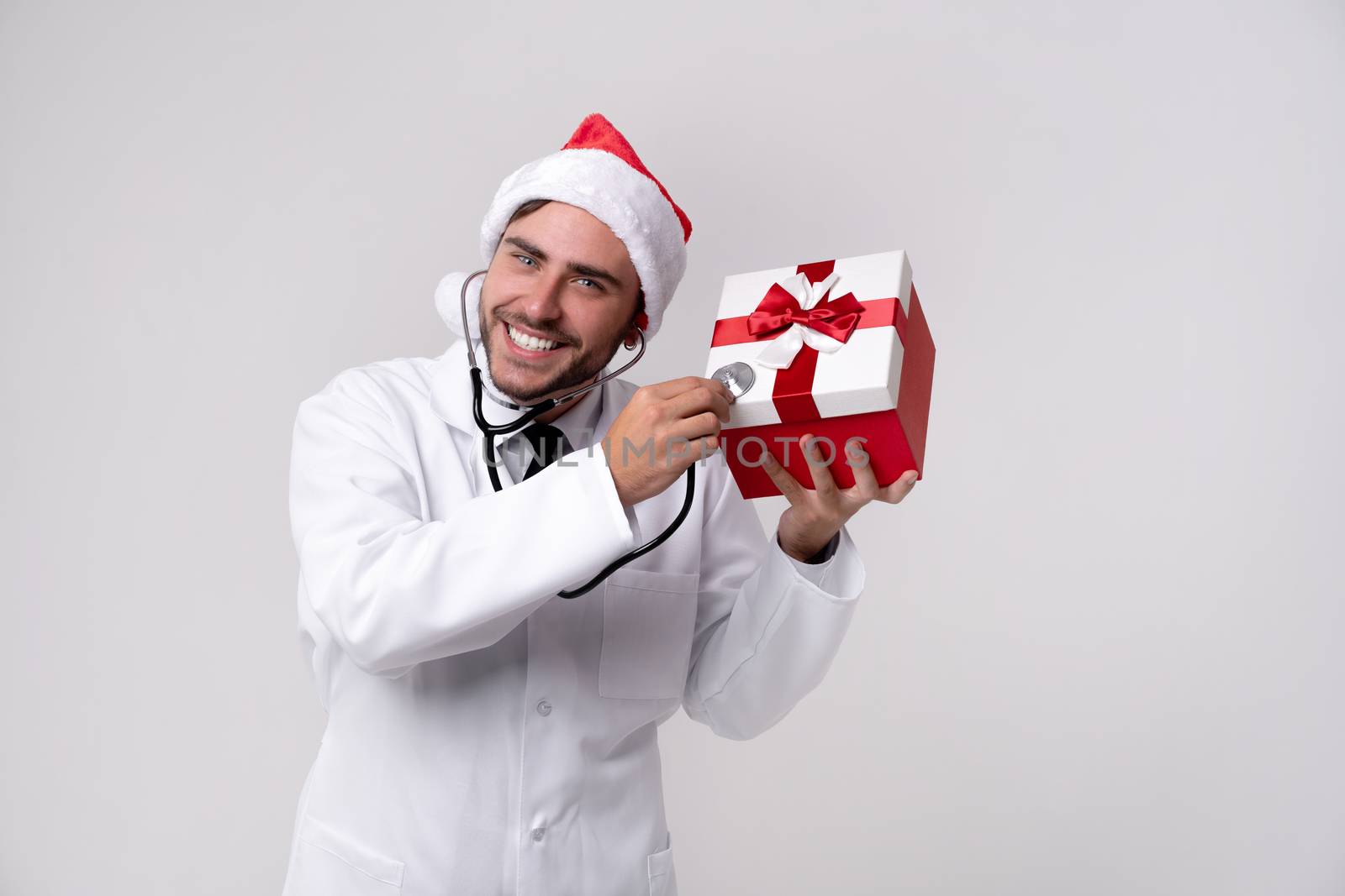 Young handsome doctor in white uniforme Santa Claus hat standing in studio on white background smile Holding red gift box in hand near ear Medical student Christmas New Year Holiday Medicine concept