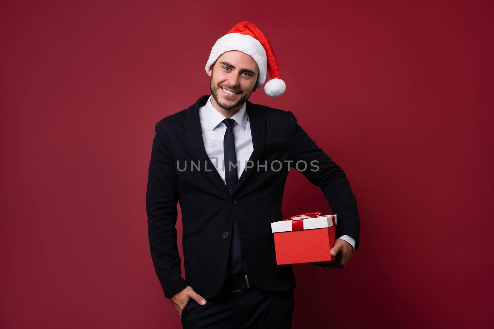 Young handsome caucasian guy in business suit and Santa hats stands on white background in studio and smilie Holding red gift box in hand. Portrait business person with Christmas mood Holiday banner