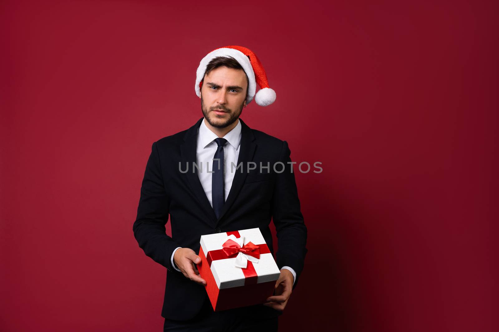 Young handsome caucasian guy in business suit and Santa hats stands on white background in studio and smilie Holding red gift box in hand. Portrait business person with Christmas mood Holiday banner