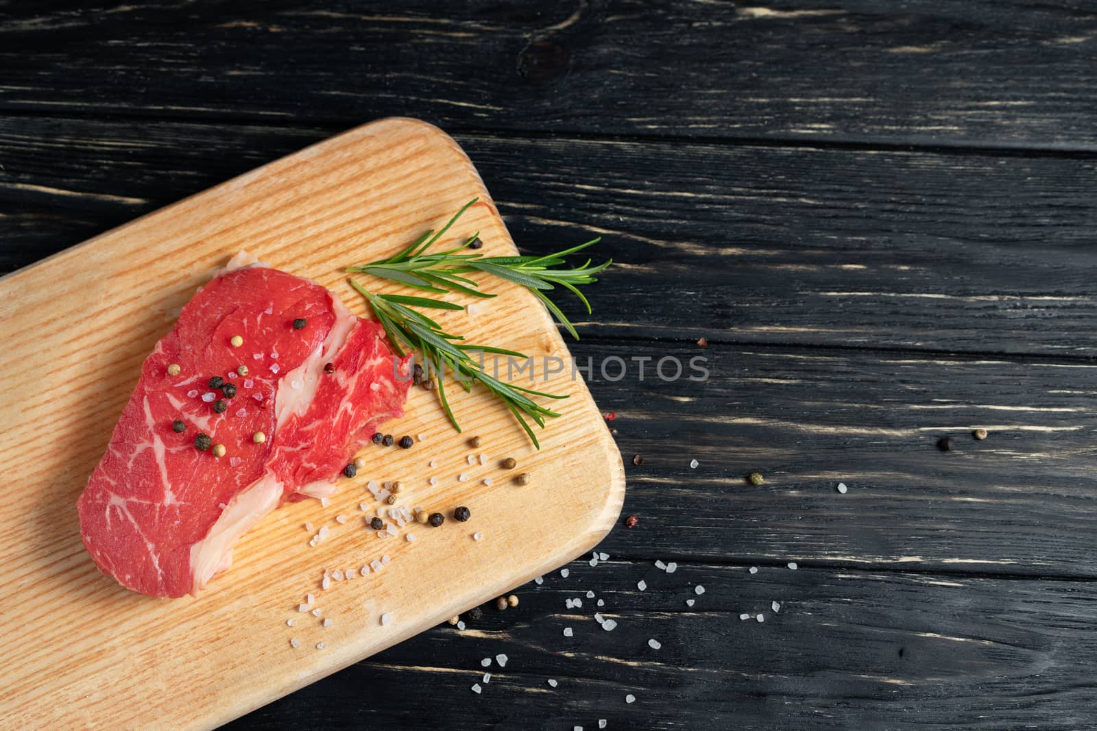one pieces of juicy raw beef with rosemary on a cutting board on a black wooden table background. Meat for barbecue or grill sprinkled with pepper and salt seasoning