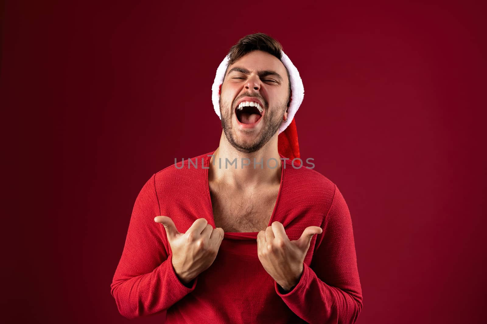 Young handsome caucasian guy in Santa hats stands on red background in studio pulls the sweater Shows how hot he is. Close up portrait european guy with Christmas mood. Holiday theme. New Year.