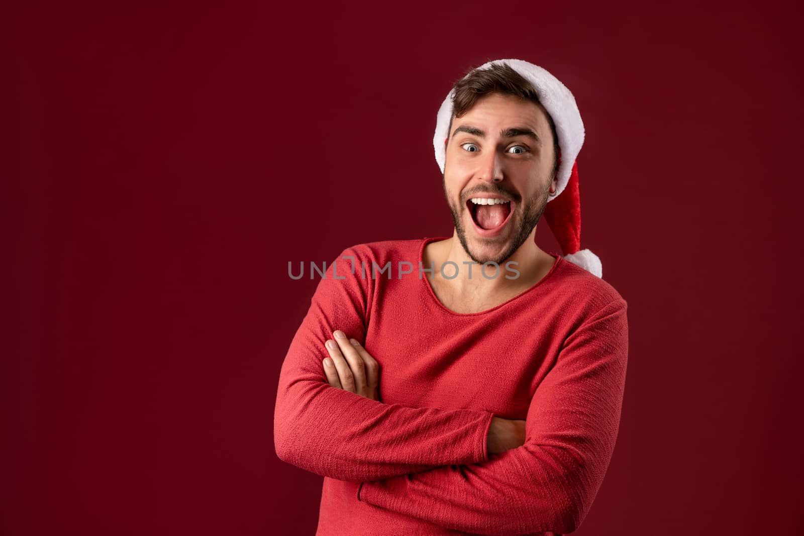 Young handsome caucasian guy in red sweater and Santa hats stands on red background in studio and teeth smiling. Close up portrait european guy with Christmas mood. He folded his arms over his chest.