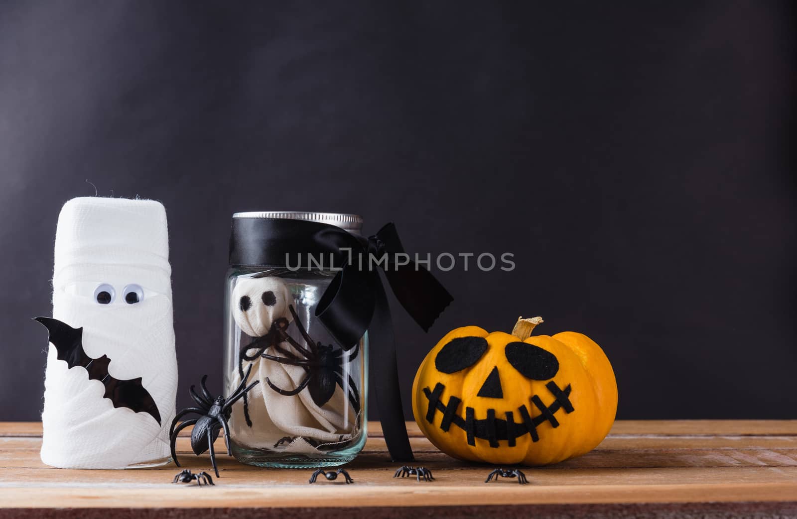 Funny Halloween day decoration party, White ghost scary face and spider in jar glass, glass of water wrapped with bandage and pumpkin on wood table, studio shot isolated on black background