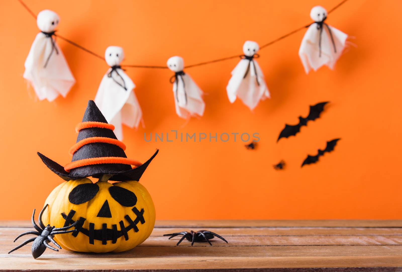 Funny Halloween day decoration party, Cute pumpkin ghost spooky jack o lantern face wear hat, black spider and bats on wooden table, studio shot isolated on an orange background, Happy holiday concept