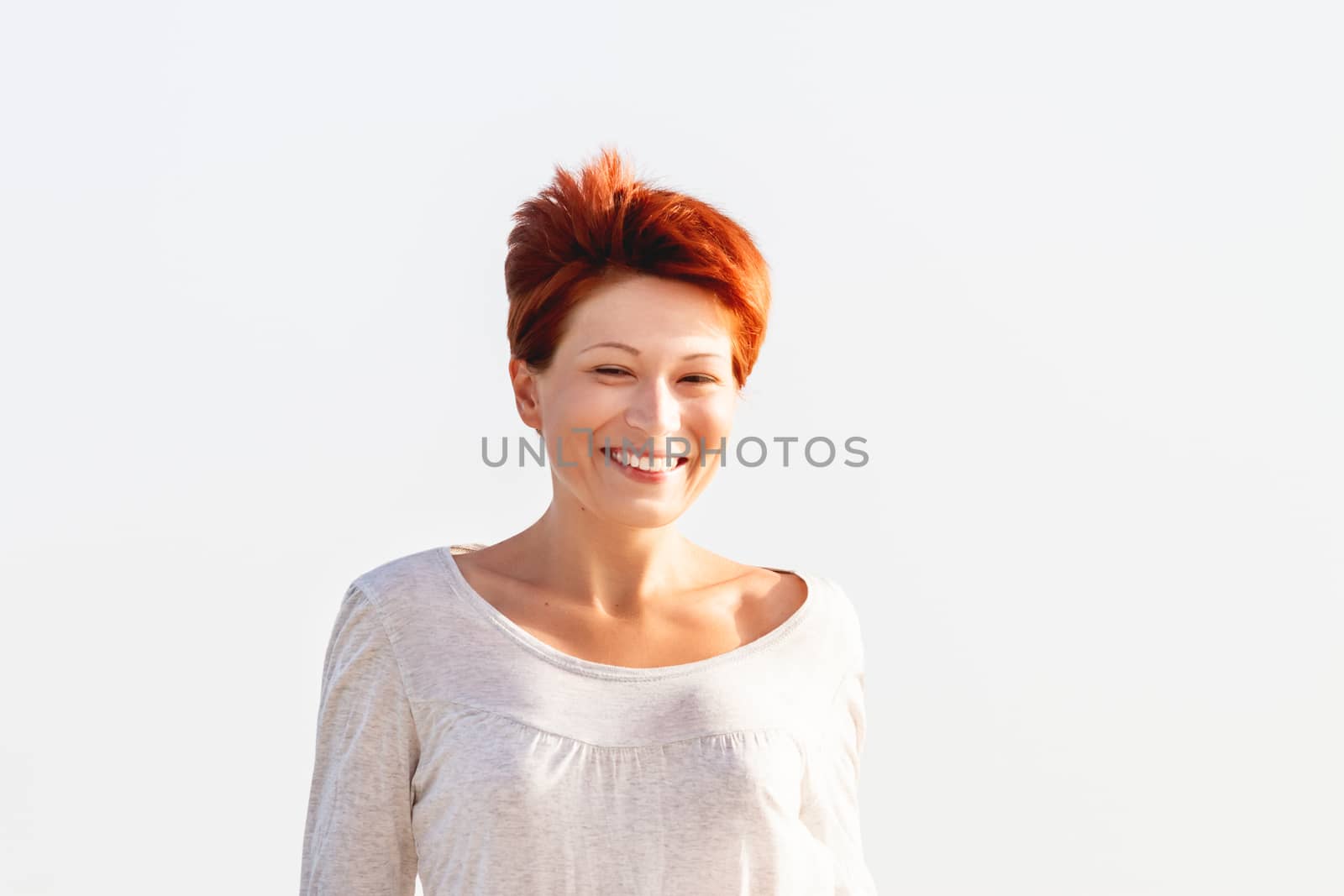 Wide smiling young woman with red short hair cut on clear sky background. Coastal morning. Natural beauty. Happiness and power of youth.
