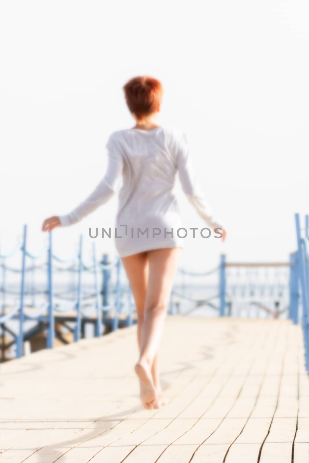 Young woman with red short hair cut running on wooden pier. Coastal morning. Natural beauty. Happiness and power of youth. Blurred background.