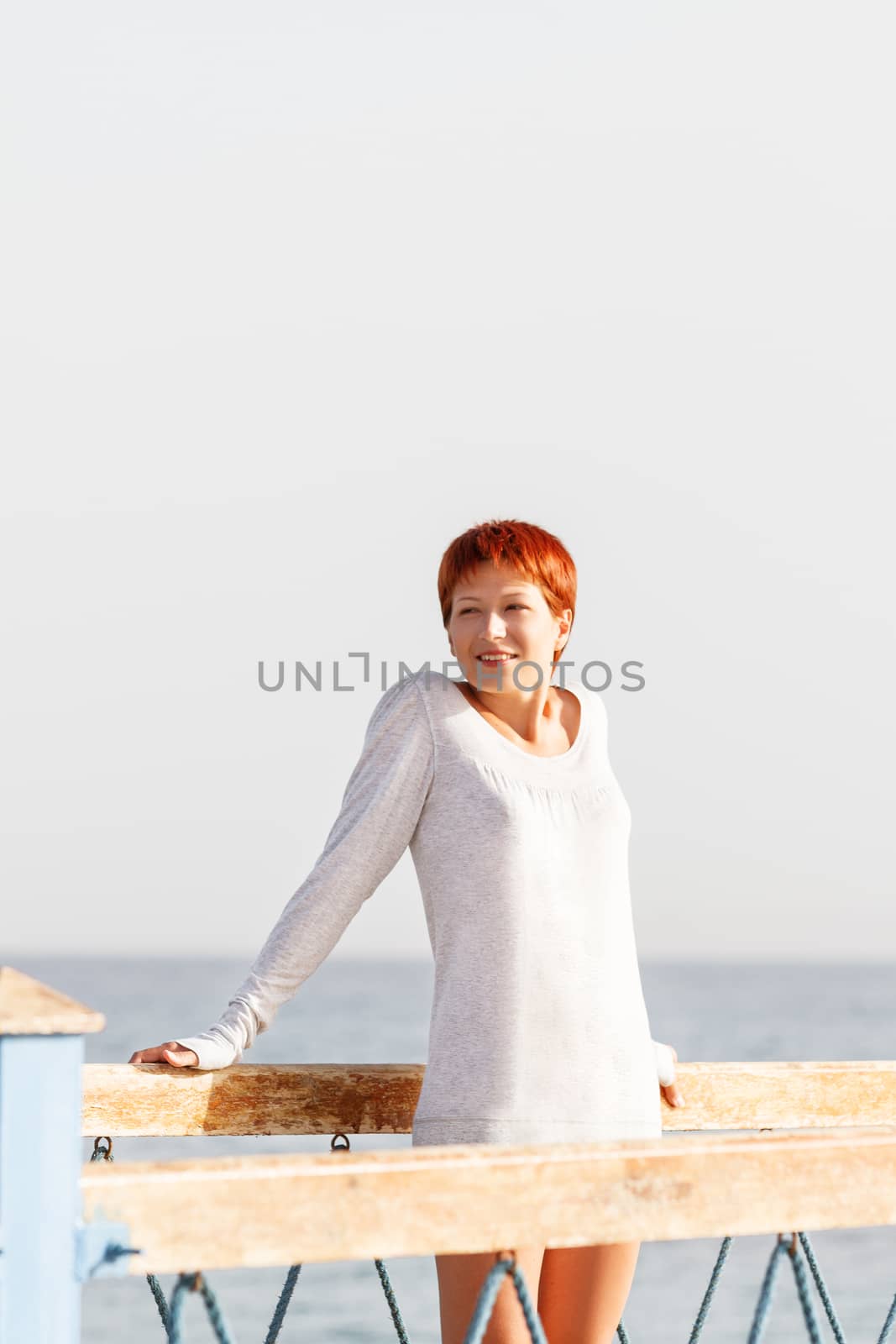 Wide smiling young woman with red short hair cut stays on wooden pier. Coastal morning sunlight. Natural beauty.