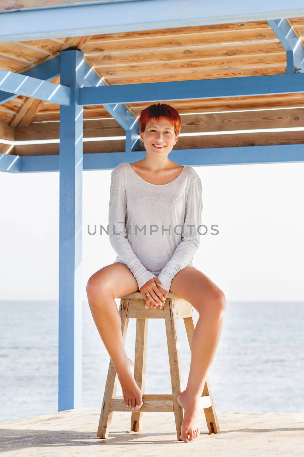 Wide smiling young woman with red short hair cut sits on wooden by aksenovko