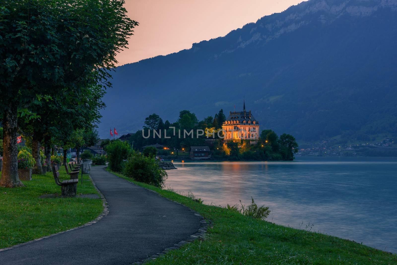 Sunset at the Iseltwald peninsula and forme castle in Switzerland, now Rehabilitation Center of Seeburg, with a public park, benches and a walkway in the foreground. Long exposure.