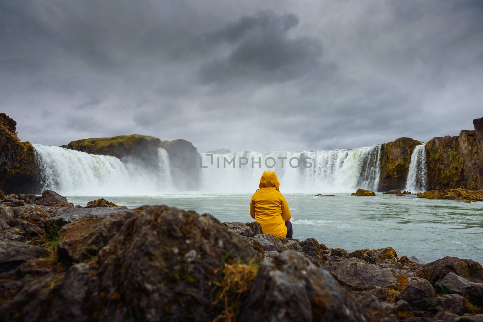 Tourist in a yellow jacket relaxing at the Godafoss waterfall in Iceland. Godafoss means the waterfall of the gods in icelandic.
