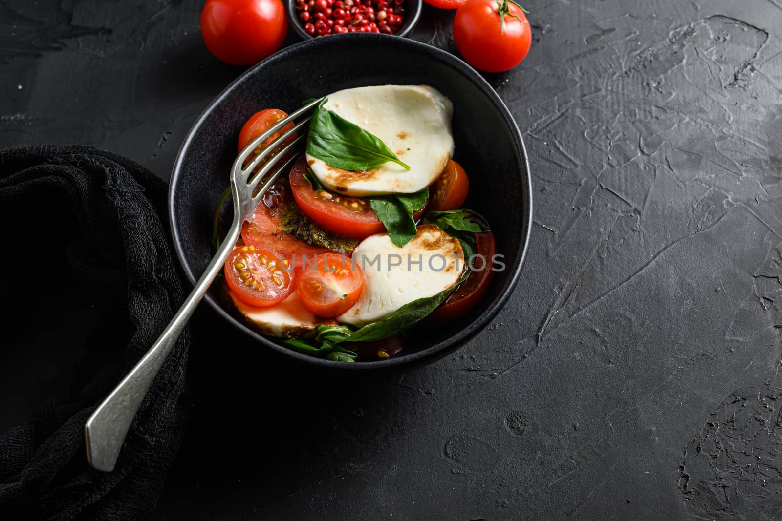 Cherry tomatoes, mozzarella buffalo cheese, basil, pesto sauce, and spices on black slate stone chalkboard. Italian traditional caprese salad ingredients. Mediterranean food in black bowl with fork space for text.