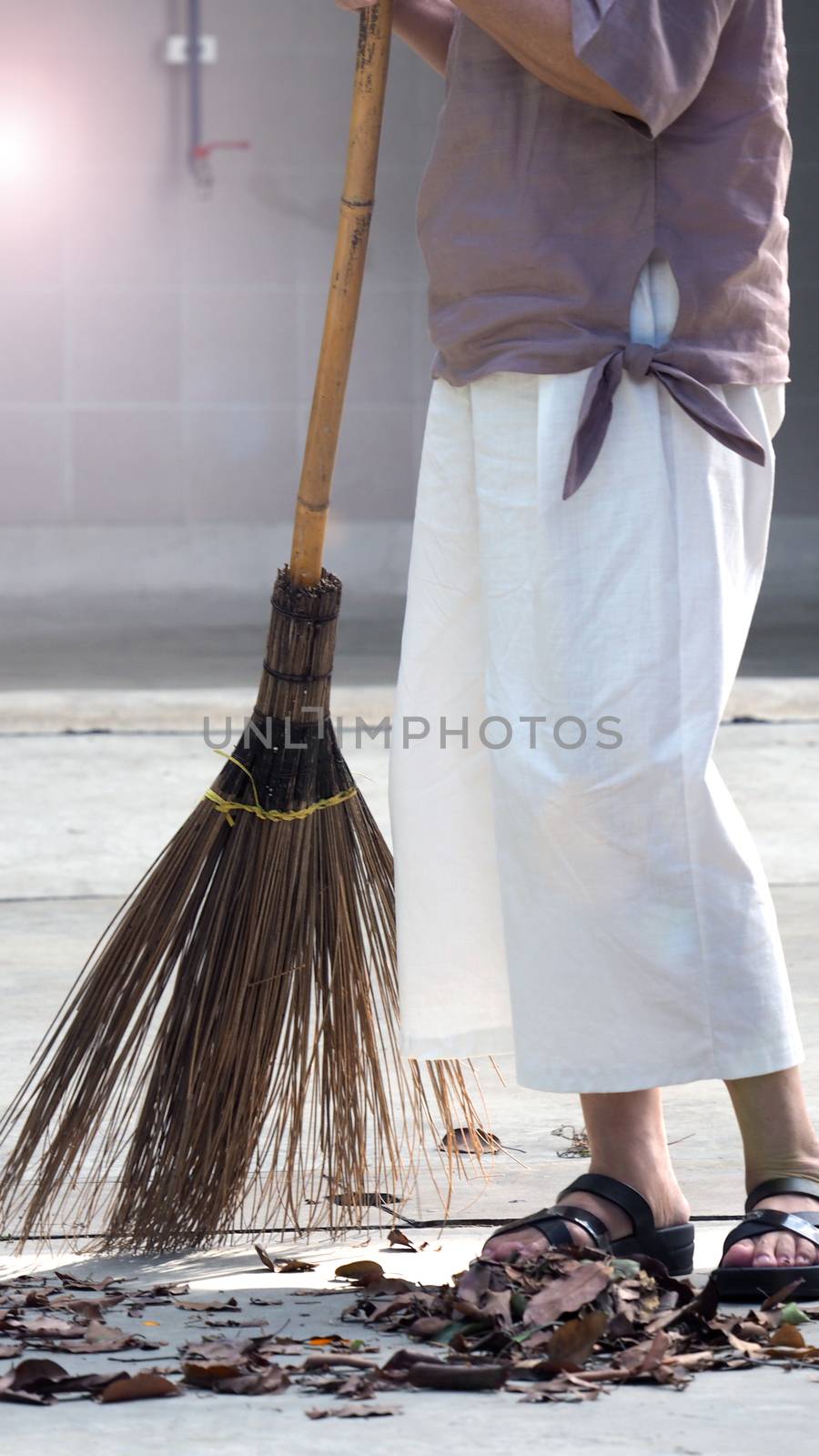 Old woman is sweeping dry leaf on the outdoor cement floor by big long broom.