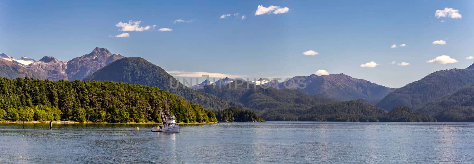 A beautiful panoramic shot of a harbour in Sitka, AK with a commercial fishing boat anchored in it, green forest, mountains with snowy peaks, and gorgeous blue sky.