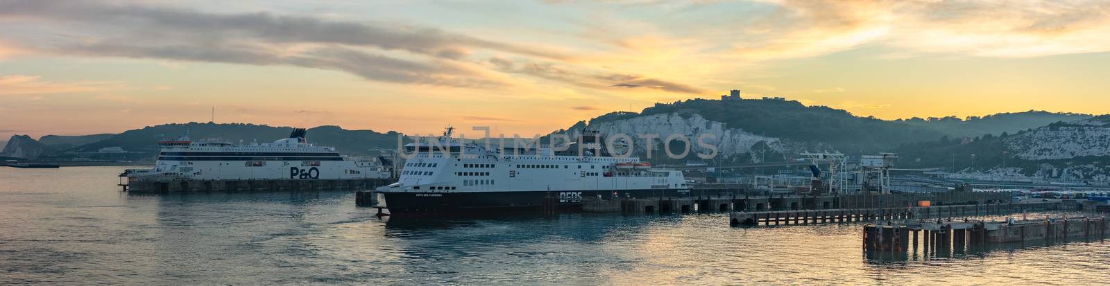 Port of Dover, England - June 24, 2020: Panoramic landscape shot of Dover Port with P&O and DFDS ferry boats docked at sunset. Beautiful warm orange sky and hills in the background.