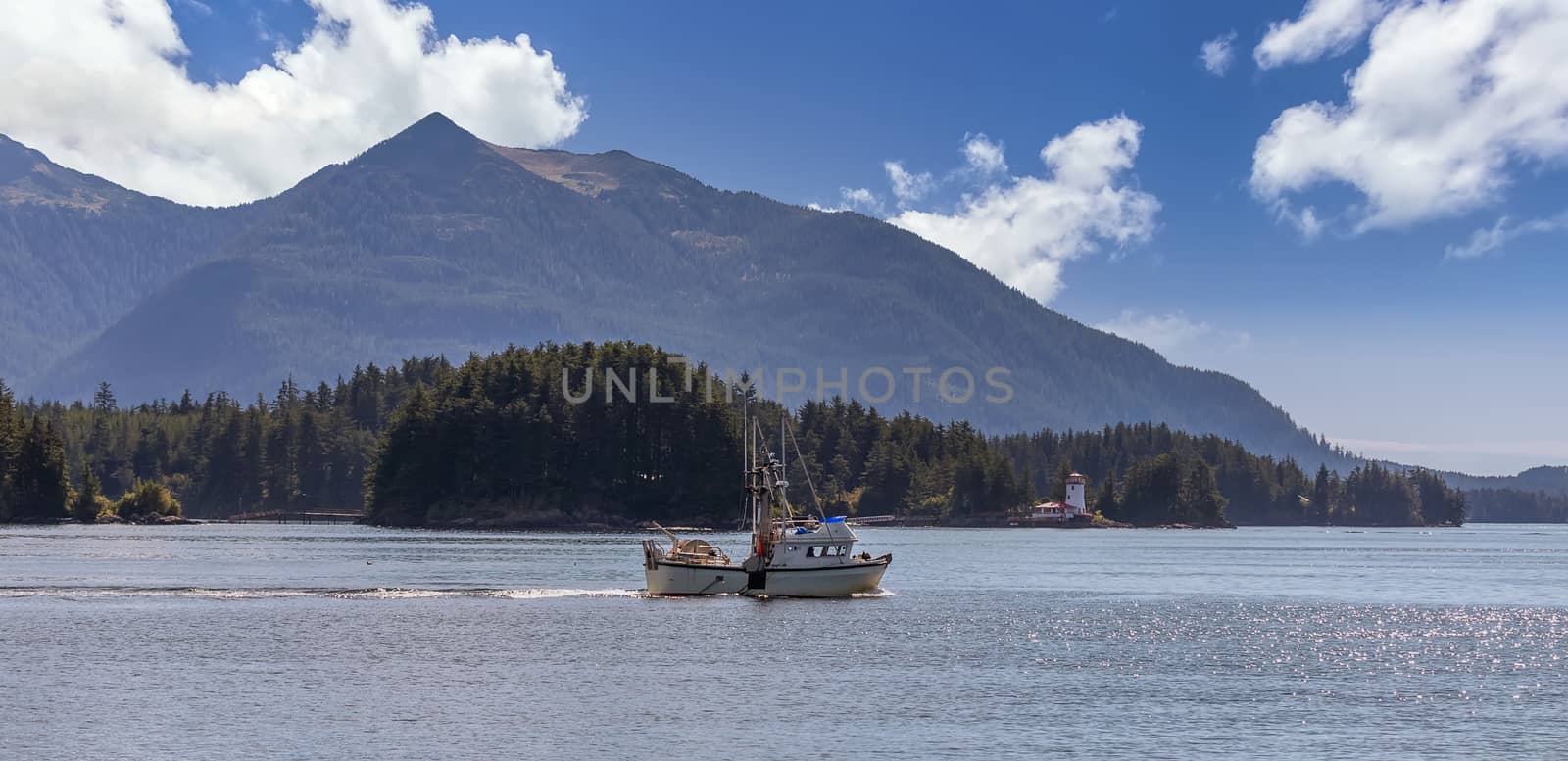 Shot of a commercial fishing boat sailing in a harbour in Sitka, AK. Green forest, mountains, beautiful blue sky with clouds, and a lighthouse in the background.