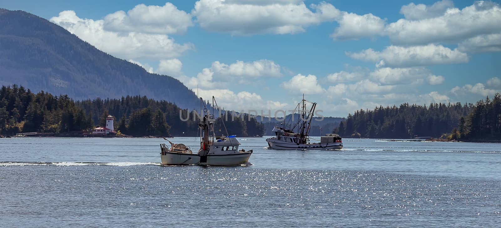 Shot of commercial fishing boats sailing in a harbour in Sitka, AK. Green forest, mountains, beautiful blue sky with clouds, and a lighthouse in the background.