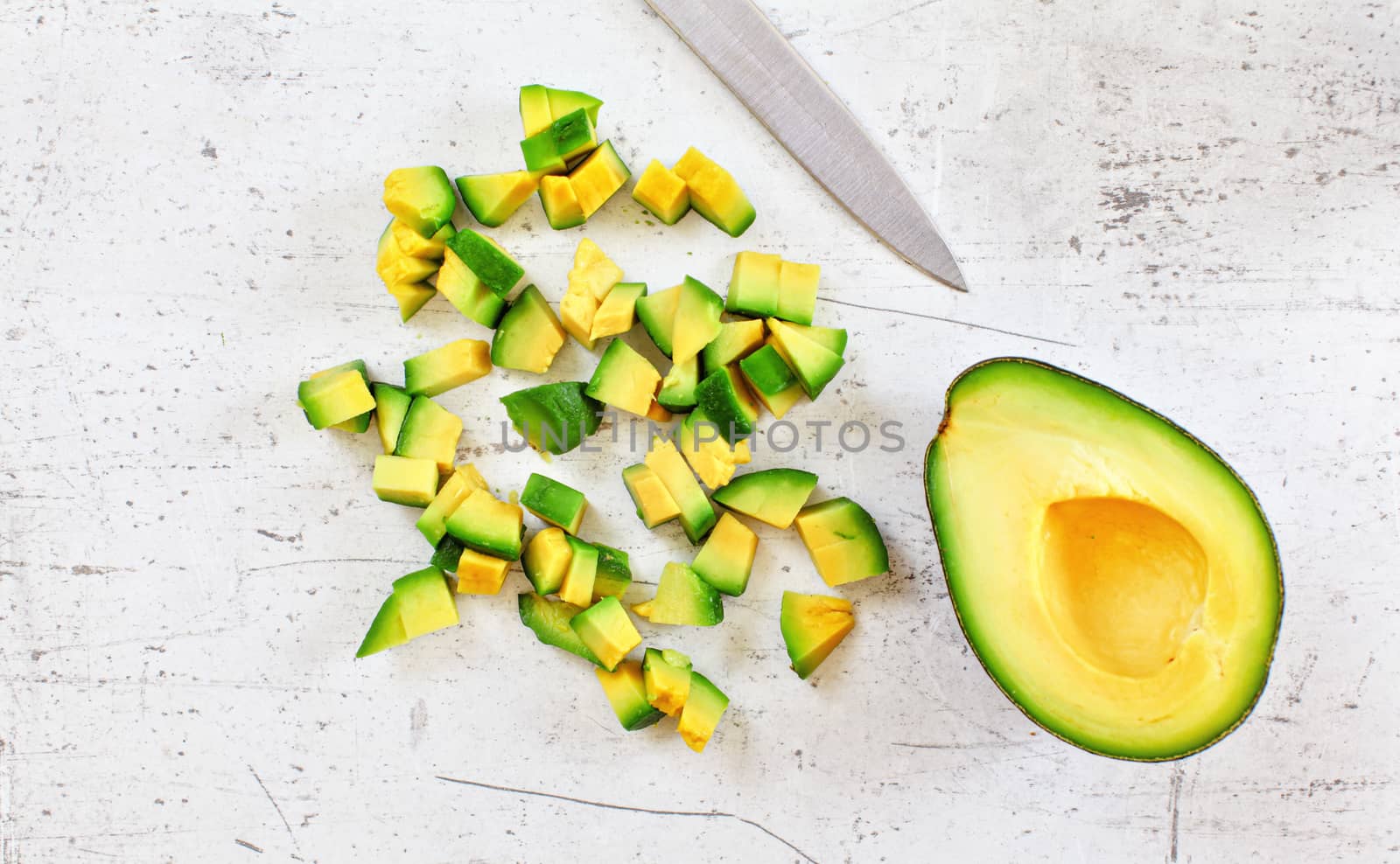 Yellow green avocado pulp cut to small pieces on white stone like desk, knife blade near, view from above by Ivanko