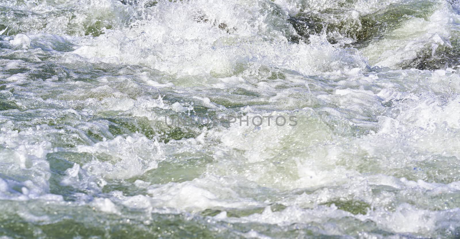 Rapid spring river flowing over rocks on sunny day, forming white water waves, closeup detail - abstract nature background.