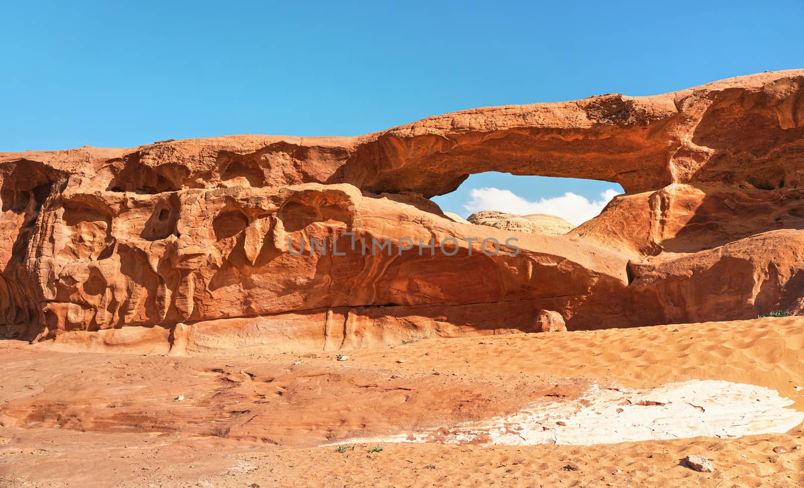 Little arc or small rock window formation in Wadi Rum desert, bright sun shines on red dust and rocks, blue sky above by Ivanko